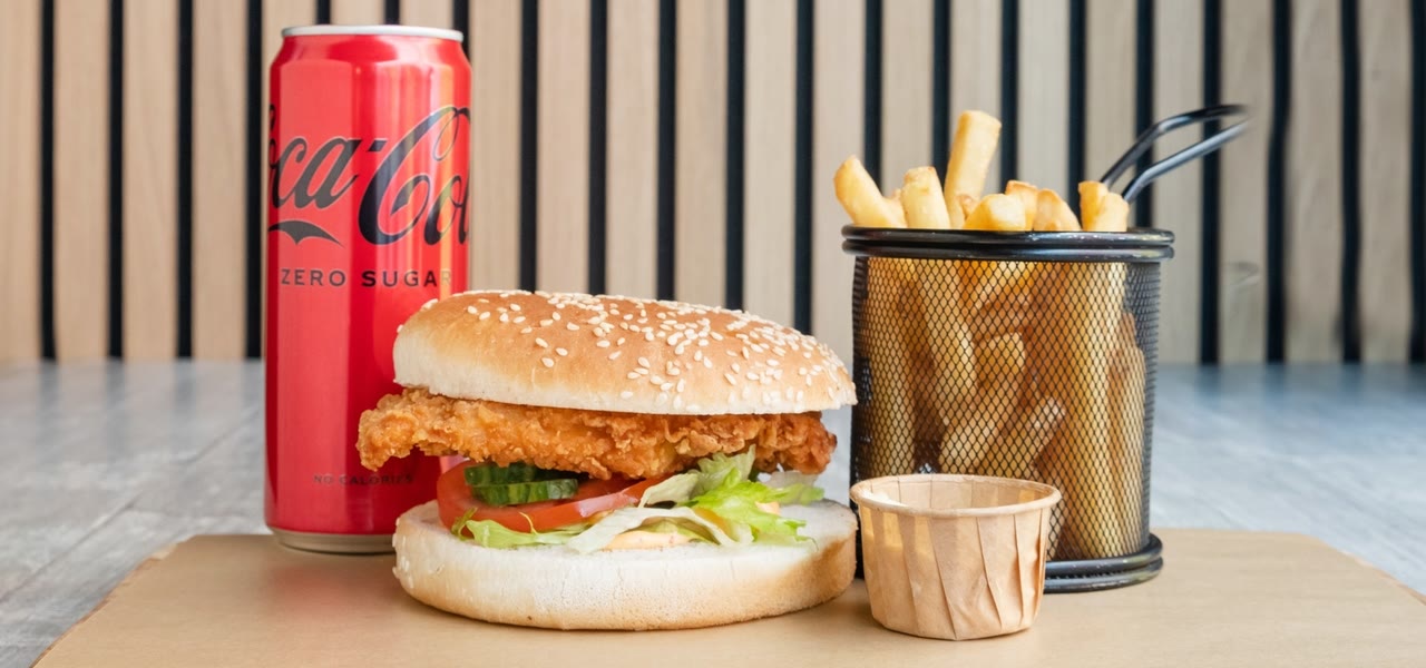 Een crispy chicken burger met friet in een mandje en een blikje cola op kraftpapier.